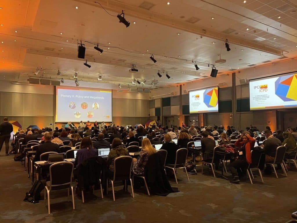 Large plenary in a conference hall, with delegates at tables and several presentation screens